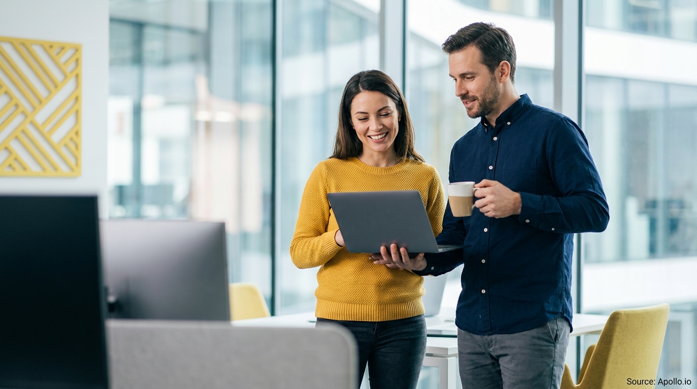 Two smiling coworkers look at a laptop screen together in a bright office.