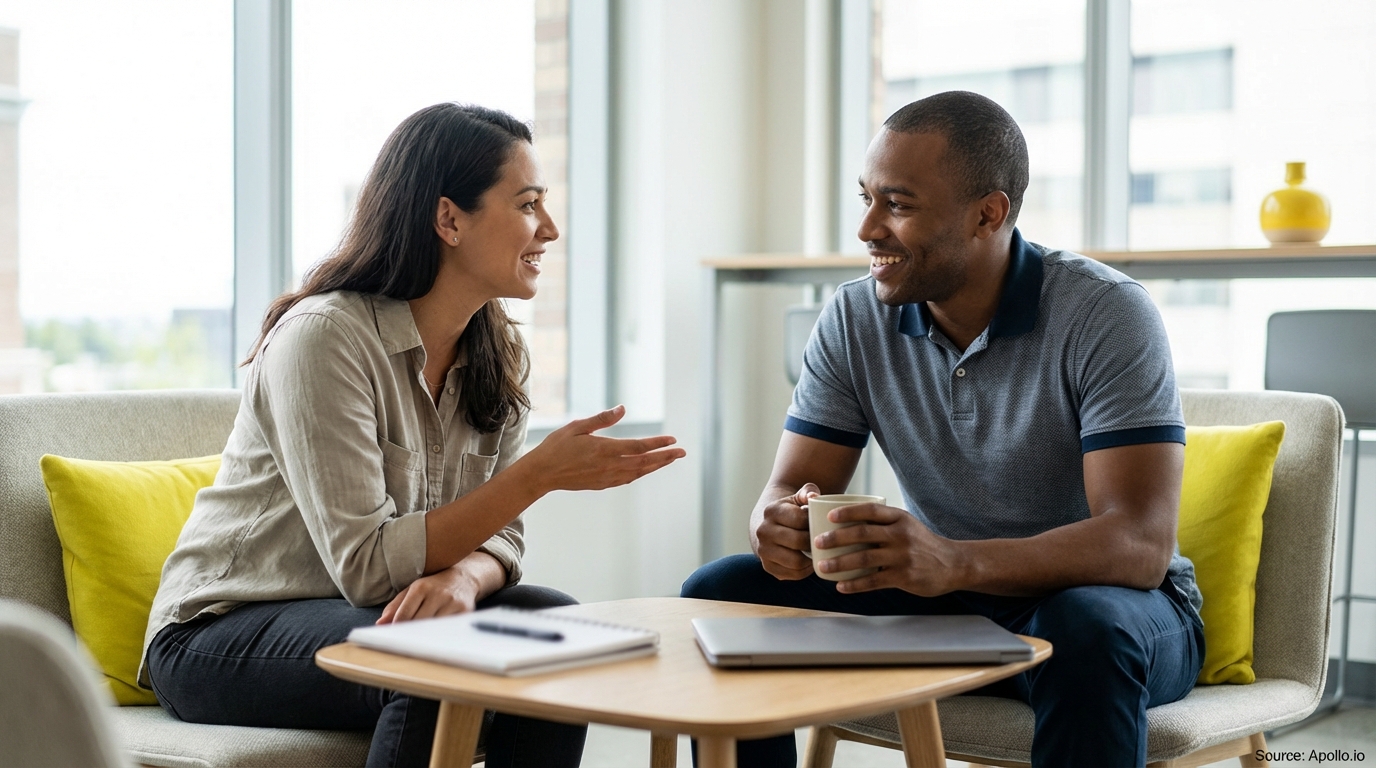 Two colleagues happily chat over coffee at a small table in a bright, modern office.