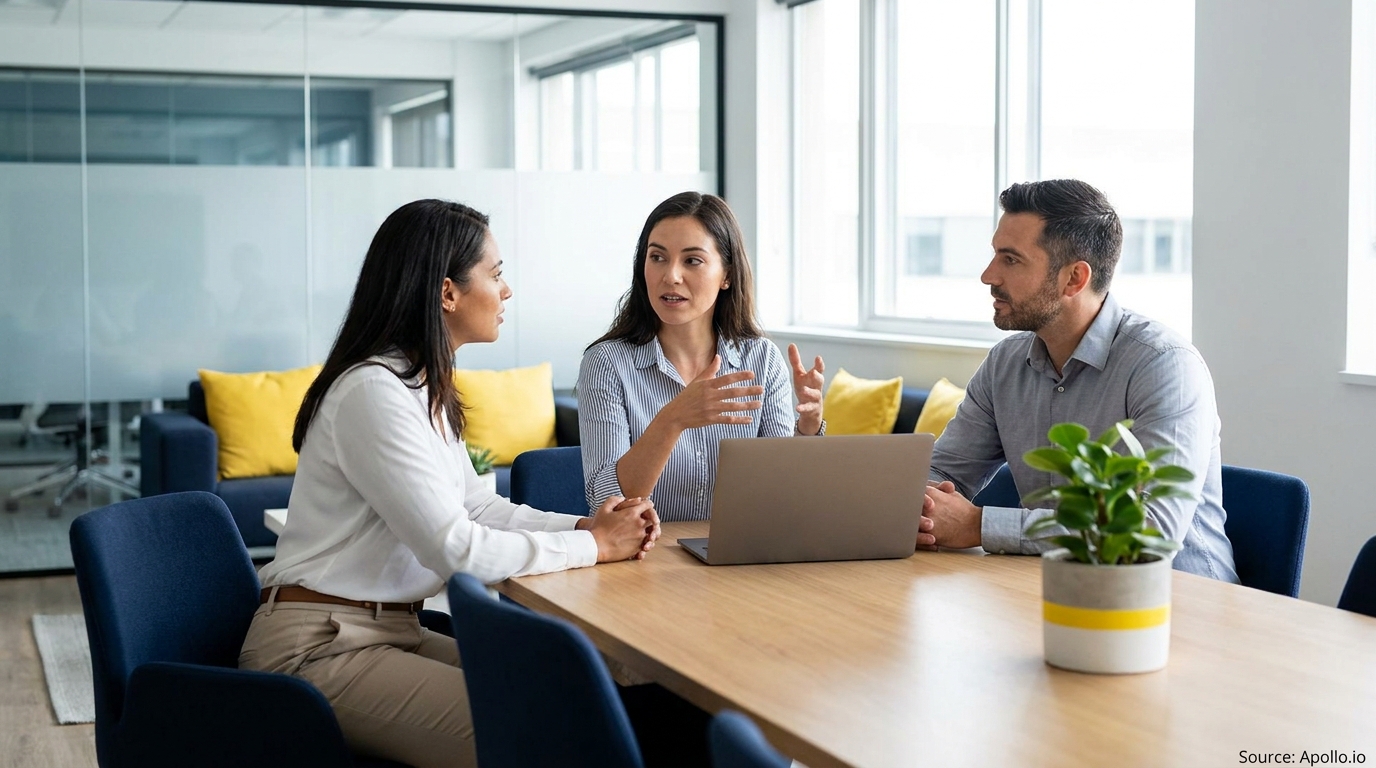 Three professionals discuss at a modern office table with a laptop.