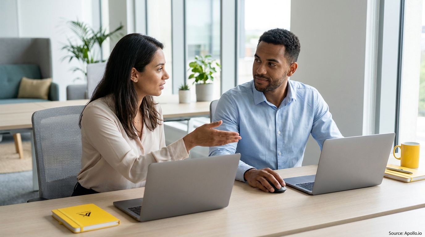 Two professionals are discussing work at a modern office desk with laptops.