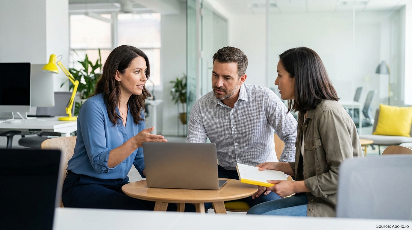 Three diverse professionals discuss work around a laptop at a modern office table.
