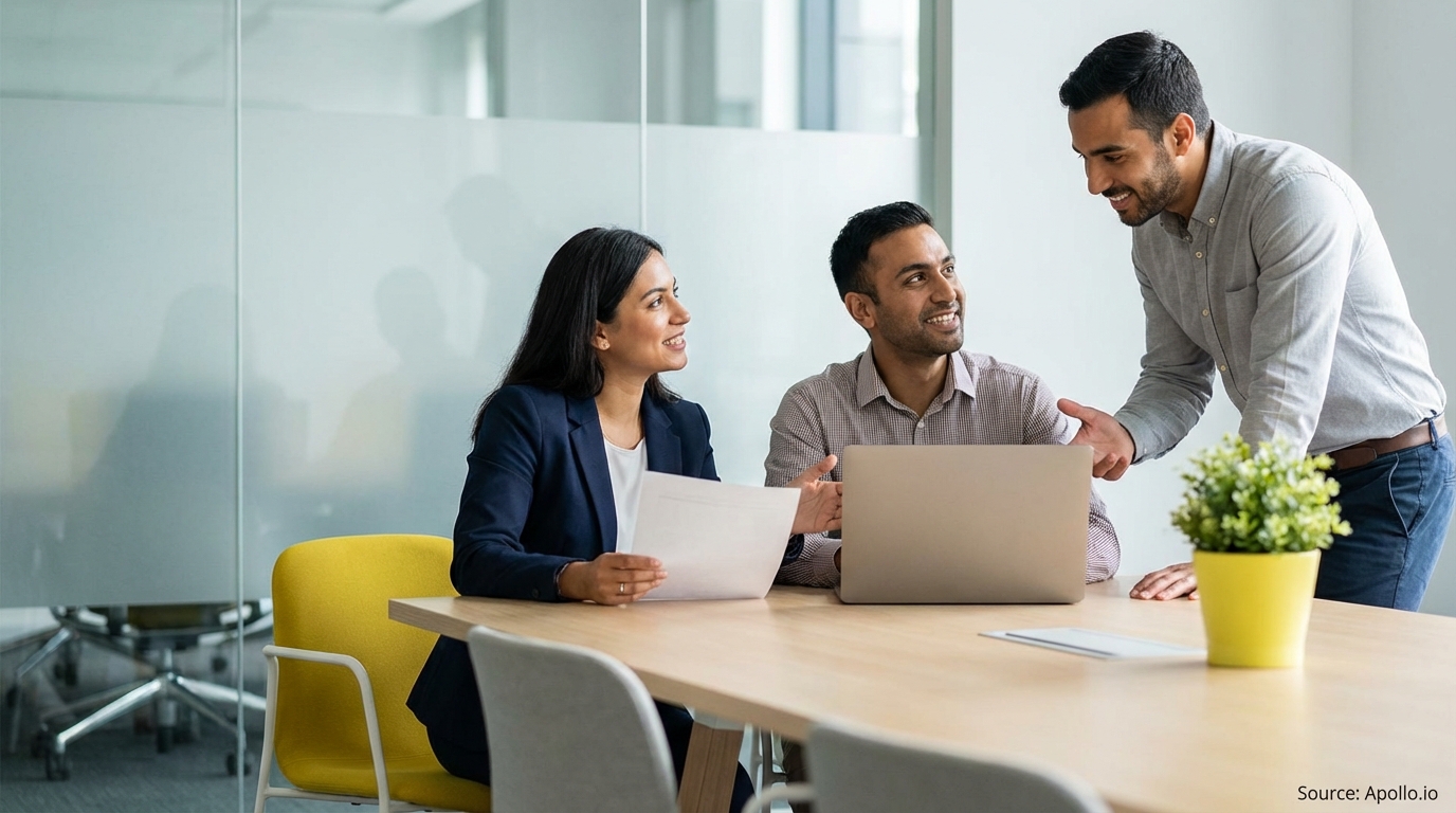 Three smiling professionals discussing work at a modern office table with a laptop.